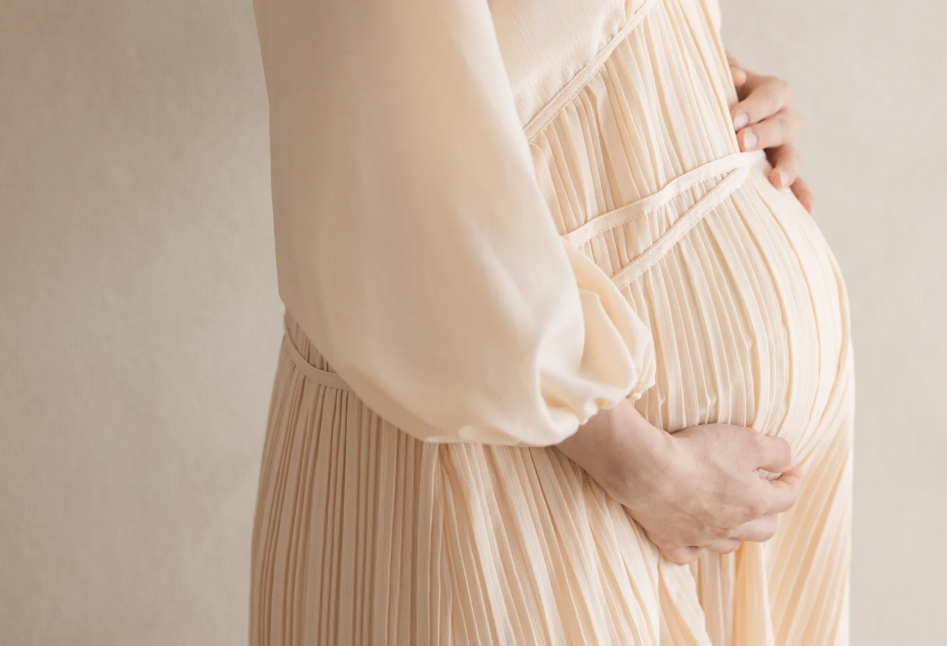 プリーツの入った薄いピンク色のドレスを着た妊婦が、大きくなったお腹を両手で優しく抱えているクローズアップ。