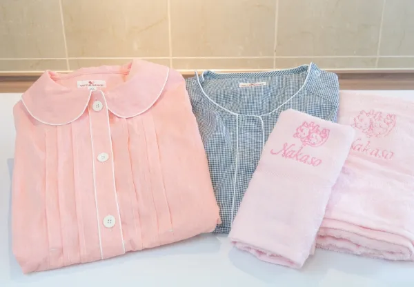 出産時、入院時に使用するアメニティグッズ。淡いピンク色の前開き分娩着、グレーの授乳口付きパジャマ、そして『Nakazawa』の刺繍が入ったピンクのタオルが並べて置かれている。