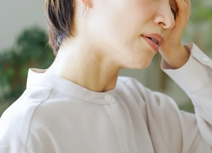 疲れた様子の女性のクローズアップ。片手で額を押さえ、首筋に力を入れている様子で、更年期障害や体調不良の症状を表している。