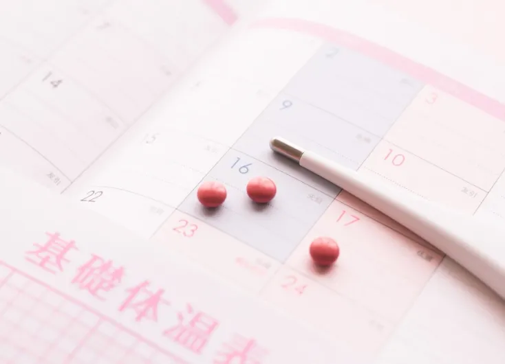ピンク色の基礎体温表の上にあるカレンダーに、赤色のピル（錠剤）が3粒置かれている。その横には体温計も見え、避妊のためのピル服用や月経周期の管理を表現している。