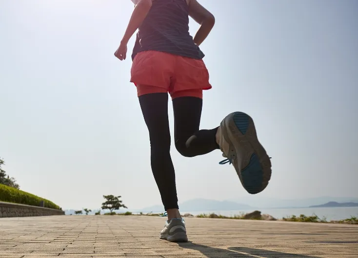 海岸沿いの遊歩道でランニングをする女性の足元。運動による健康維持や生活習慣の改善を表現している。